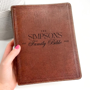 May include: A brown leather-bound Bible with the words "THE SIMPSONS Family Bible 2025" printed on the cover. The cover has a textured appearance and a stitched border. The word "EST." is printed on the left side of the cover.