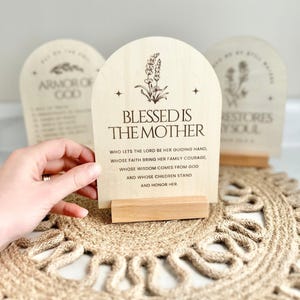 May include: Wooden sign with a floral design and the text "Blessed is the Mother" followed by a quote about a mother's guiding hand, faith, wisdom, and honor.