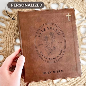 May include: A brown leather Holy Bible with a gold cross and personalized text. The cover features an oval design with the name "Elizabeth", floral illustrations, and the text "Daughter of a King". The words "Holy Bible" are printed at the bottom.