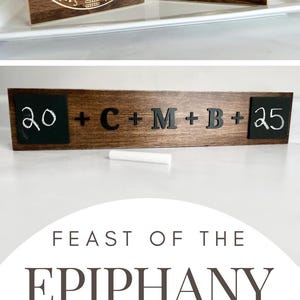 May include: A wooden sign with a chalkboard insert that reads "20 + C + M + B + 25". The sign is decorated with a brown stain and has a piece of white chalk next to it. The sign is advertising a guide for the Feast of the Epiphany.