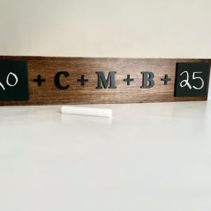 May include: A wooden sign with two chalkboard sections, one with the number 20 written in white chalk and the other with the number 25 written in white chalk. The middle section of the sign has the letters C, M, and B written in black, with plus signs between each letter.
