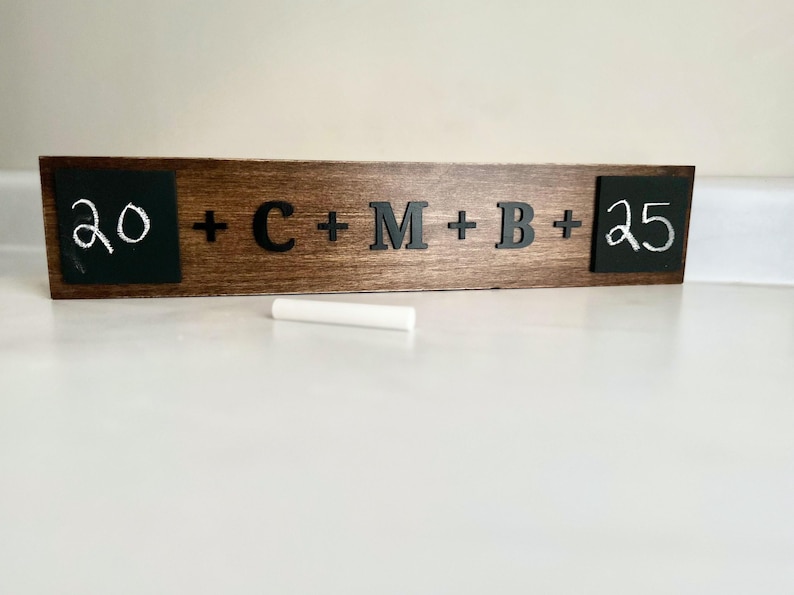 May include: A wooden sign with two chalkboard sections, one with the number 20 written in white chalk and the other with the number 25 written in white chalk. The middle section of the sign has the letters C, M, and B written in black, with plus signs between each letter.