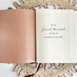 K&ouml;nnte beinhalten: Ge&ouml;ffnete Bibel mit dem Text "NIV Journal the Word Bible Double-Column". Die linke Seite hat ein braunes Punktmuster, w&auml;hrend die rechte Seite wei&szlig; ist. Die Bibel liegt auf einer beigefarbenen, geh&auml;kelten Tischdecke.