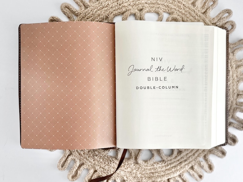 K&ouml;nnte beinhalten: Ge&ouml;ffnete Bibel mit dem Text "NIV Journal the Word Bible Double-Column". Die linke Seite hat ein braunes Punktmuster, w&auml;hrend die rechte Seite wei&szlig; ist. Die Bibel liegt auf einer beigefarbenen, geh&auml;kelten Tischdecke.