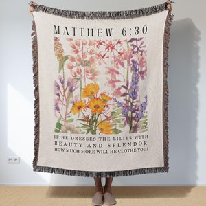 May include: A woven throw blanket with a floral design and the text "MATTHEW 6:30" and "IF HE DRESSES THE LILIES WITH BEAUTY AND SPLENDOR HOW MUCH MORE WILL HE CLOTHE YOU?"
