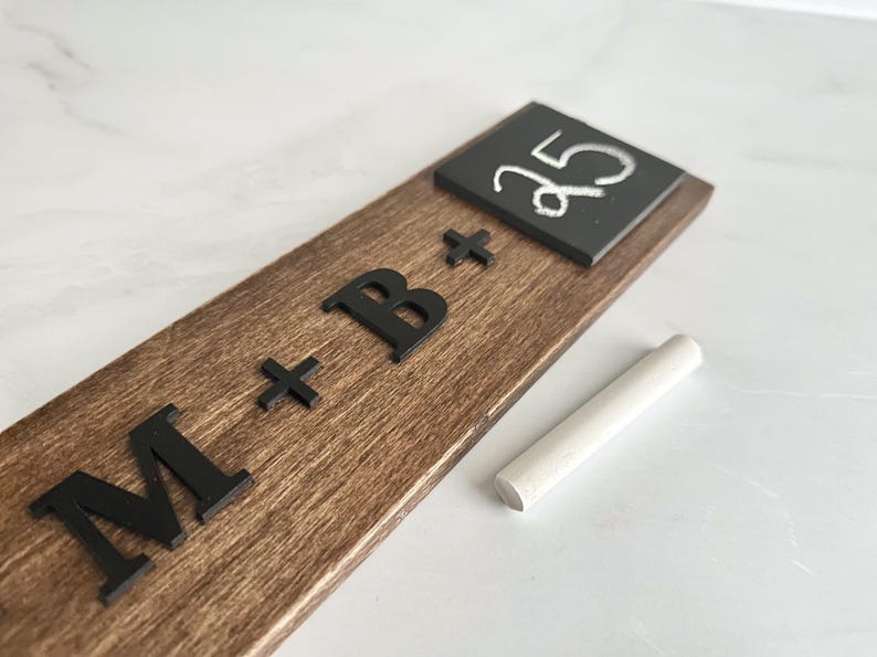 May include: A wooden sign with the text "M + B +" and a small chalkboard with the number "35" written on it. A piece of chalk is lying next to the sign.