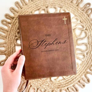 May include: A brown leather-bound book with the name "Stephens" and the year "2026" embossed on the cover. A gold cross is in the upper right corner. The book is on a beige woven placemat.