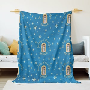 May include: A blue fleece blanket with a repeating pattern of Our Lady of Guadalupe images and the text "Our Lady of Guadalupe Pray For Us" in gold lettering. The background is a blue sky with white stars.