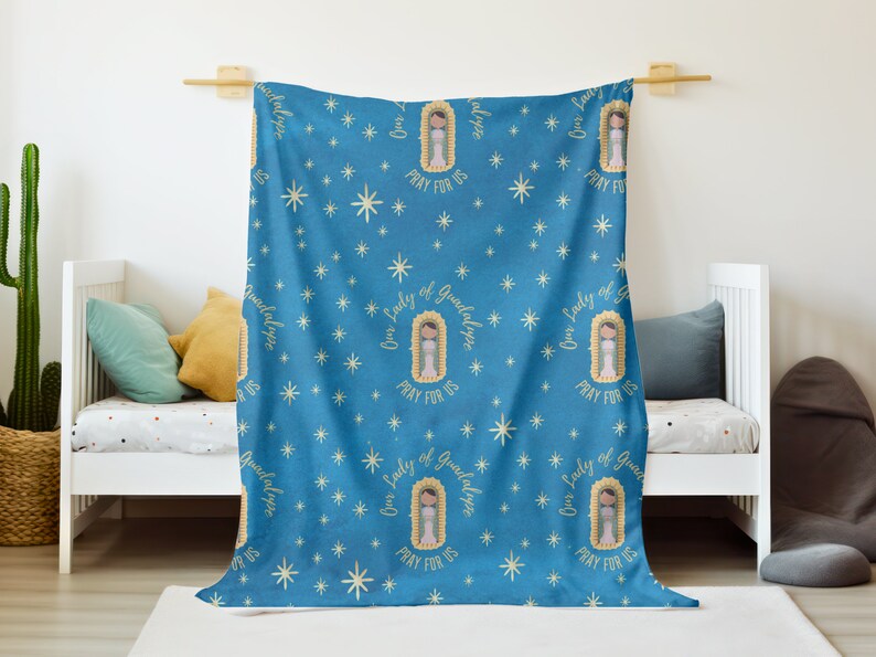 May include: A blue fleece blanket with a repeating pattern of Our Lady of Guadalupe images and the text "Our Lady of Guadalupe Pray For Us" in gold lettering. The background is a blue sky with white stars.