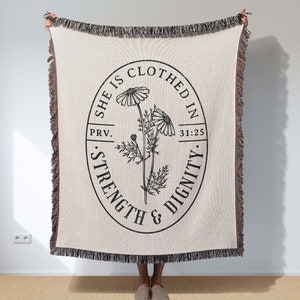 May include: A woven throw blanket with a cream background and a black circle design. The circle features a floral illustration and the text "She is clothed in strength & dignity. Prov. 31:25."