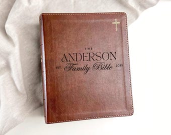 May include: A brown leather Family Bible with the text "The Anderson Family Bible" and the year 2025. A gold cross is on the upper right corner. The Bible is on a white surface.