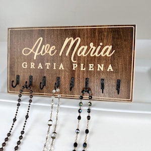 May include: A wooden jewelry organizer with the text "Ave Maria Gratia Plena" in gold lettering. The organizer has eight hooks for hanging necklaces and bracelets.