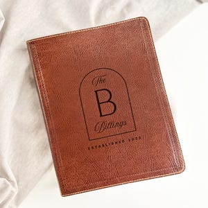 May include: A brown leather journal with the text "The B Billings ESTABLISHED 2025" in a black arched design. The journal is rectangular and has a textured surface. The journal is resting on a light-colored fabric.