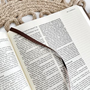 K&ouml;nnte beinhalten: Ein offenes Buch mit einem braunen Leseband und einer strukturierten beigefarbenen Oberfl&auml;che. Die Seite zeigt Text aus einem religi&ouml;sen Text, wobei das Leseband zwischen den Seiten liegt.