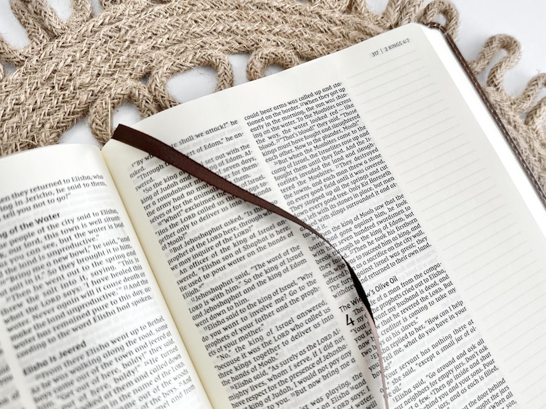 K&ouml;nnte beinhalten: Ein offenes Buch mit einem braunen Leseband und einer strukturierten beigefarbenen Oberfl&auml;che. Die Seite zeigt Text aus einem religi&ouml;sen Text, wobei das Leseband zwischen den Seiten liegt.