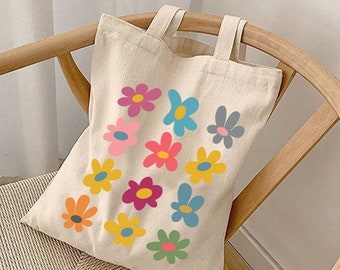 May include: A white canvas tote bag with colorful flower designs. The bag has two handles and is sitting on a wooden chair.