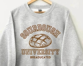 Puede incluir: Una sudadera gris con el texto "SOURDOUGH UNIVERSITY BREADUCATED" en letras marrones. Un dibujo de una hogaza de pan de masa madre está por encima del texto.