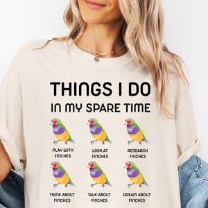 May include: Cream-colored t-shirt with the text "THINGS I DO IN MY SPARE TIME" above six colorful finch illustrations. Each finch illustration is paired with a phrase: "PLAY WITH FINCHES", "LOOK AT FINCHES", "RESEARCH FINCHES", "THINK ABOUT FINCHES", "TALK ABOUT FINCHES", and "DREAM ABOUT FINCHES".