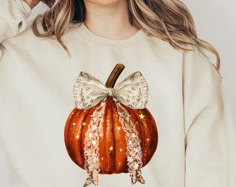 May include: Beige sweatshirt featuring a vibrant orange pumpkin graphic with a decorative bow. The pumpkin has a textured appearance with highlights. The sweatshirt is long-sleeved and has a crew neck.