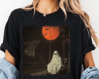 May include: Black t-shirt featuring a vintage-style Halloween graphic. The design depicts a ghost on a swing set in front of a house, with a large orange moon and bats in the sky. The shirt is a comfortable, casual style.