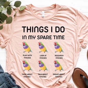 May include: A light pink t-shirt with the text "THINGS I DO IN MY SPARE TIME" and finch graphics. The shirt also has the phrases "PLAY WITH FINCHES", "LOOK AT FINCHES", "RESEARCH FINCHES", "THINK ABOUT FINCHES", "TALK ABOUT FINCHES", and "DREAM ABOUT FINCHES".