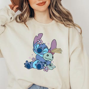 K&ouml;nnte beinhalten: Cremefarbenes Sweatshirt mit einer Cartoonfigur in Blau-, Lila- und Rosat&ouml;nen. Die Grafik zeigt die Figur, die eine Puppe umarmt, umgeben von Blumen. Das Sweatshirt hat lange &Auml;rmel und einen Rundhalsausschnitt.