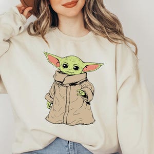 May include: A cream colored sweatshirt with a green cartoon character wearing a brown coat. The character has large ears and is looking to the left. The sweatshirt has a brown hat on top of it.