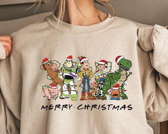 Può includere: Una felpa beige con un design natalizio con personaggi del franchise Toy Story. I personaggi indossano cappelli da Babbo Natale e sono circondati da luci natalizie. Il testo "Merry Christmas" è stampato sotto i personaggi.
