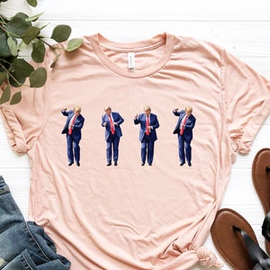 May include: A light pink t-shirt featuring a repeating image of a person in a blue suit and red tie. The shirt is laid flat with a pair of blue jeans and black sandals. Green leafy sprigs are in the upper left corner.