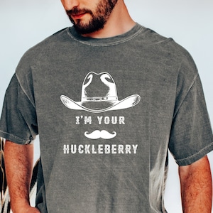 May include: A gray t-shirt with the text "I'M YOUR HUCKLEBERRY" in white, along with a cowboy hat and mustache graphic. The shirt is a relaxed fit and has short sleeves.