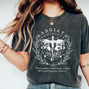 Peut inclure: Un t-shirt gris fonc&eacute; avec un motif blanc repr&eacute;sentant un oiseau aux ailes d&eacute;ploy&eacute;es, entour&eacute; de feuilles, de lunes et d'&eacute;toiles. Le texte "BASGIATH WAR COLLEGE" est &eacute;crit en cercle autour de l'oiseau. Le texte "There's nowhere in existence you could go that I wouldn't find you, Holence" est &eacute;crit sous le motif.