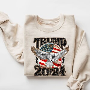 May include: A beige sweatshirt with a black graphic print of an eagle flying over an American flag with the text "TRUMP 2024".