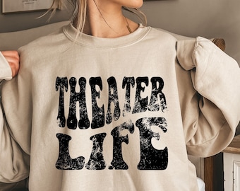 May include: A beige sweatshirt with the words "THEATER LIFE" printed in distressed black lettering. The sweatshirt has a crew neck and long sleeves. The image is of a person wearing the sweatshirt.