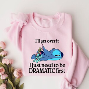 May include: Pink sweatshirt with a blue Stitch character from Lilo & Stitch. The text on the sweatshirt reads "I'll get over it I just need to be DRAMATIC first."