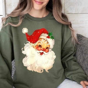 May include: A green sweatshirt with a vintage illustration of Santa Claus's face. The illustration features a red hat with white trim and a white beard.