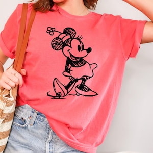 May include: A coral-colored t-shirt featuring a black graphic of Minnie Mouse. The cartoon character is depicted in a classic pose, wearing a skirt and shoes. The shirt has short sleeves and a relaxed fit, suitable for casual wear.