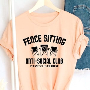 May include: Peach coloured t-shirt with black text that reads "Fence Sitting Anti-Social Club Please Sit Over There". The text is surrounded by a graphic of three chairs.