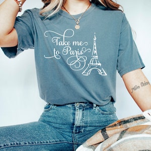 May include: A gray t-shirt with the text "Take me to Paris" and a white outline of the Eiffel Tower. The shirt is being worn by a person with long blonde hair and a gold necklace.