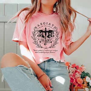Peut inclure: Un t-shirt rose avec un motif graphique noir repr&eacute;sentant une cr&eacute;ature ail&eacute;e, une couronne et le texte "BASGIATH WAR COLLEGE". Le texte "There's nowhere in existence you could go that I wouldn't find you, Valance." est imprim&eacute; sous le graphique.