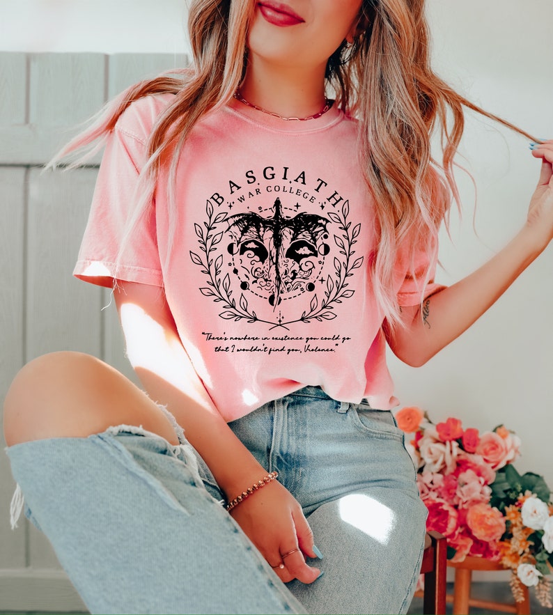 Peut inclure: Un t-shirt rose avec un motif graphique noir repr&eacute;sentant une cr&eacute;ature ail&eacute;e, une couronne et le texte "BASGIATH WAR COLLEGE". Le texte "There's nowhere in existence you could go that I wouldn't find you, Valance." est imprim&eacute; sous le graphique.