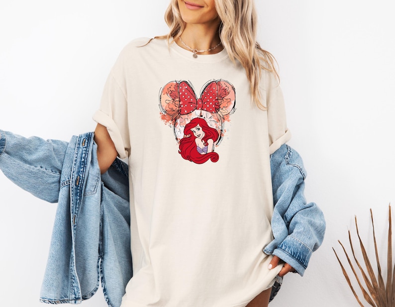 May include: A white t-shirt with a graphic of Ariel from The Little Mermaid in a red bow, with a watercolor-style background.