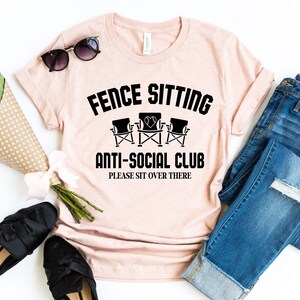 May include: A pink t-shirt with the text "Fence Sitting Anti-Social Club Please Sit Over There" and a graphic of three chairs. The shirt is laid out on a white surface with a pair of jeans, sunglasses, and a bouquet of flowers.