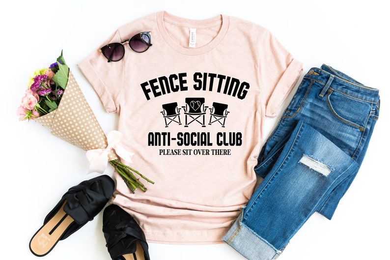 May include: A pink t-shirt with the text "Fence Sitting Anti-Social Club Please Sit Over There" and a graphic of three chairs. The shirt is laid out on a white surface with a pair of jeans, sunglasses, and a bouquet of flowers.