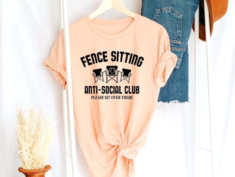 May include: Peach coloured t-shirt with black text that reads "Fence Sitting Anti-Social Club Please Sit Over There". The text is surrounded by a graphic of three chairs.