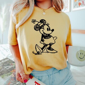 May include: Yellow t-shirt featuring a black and white illustration of Minnie Mouse. The shirt is paired with light blue shorts. A white mug with a smiley face is visible.