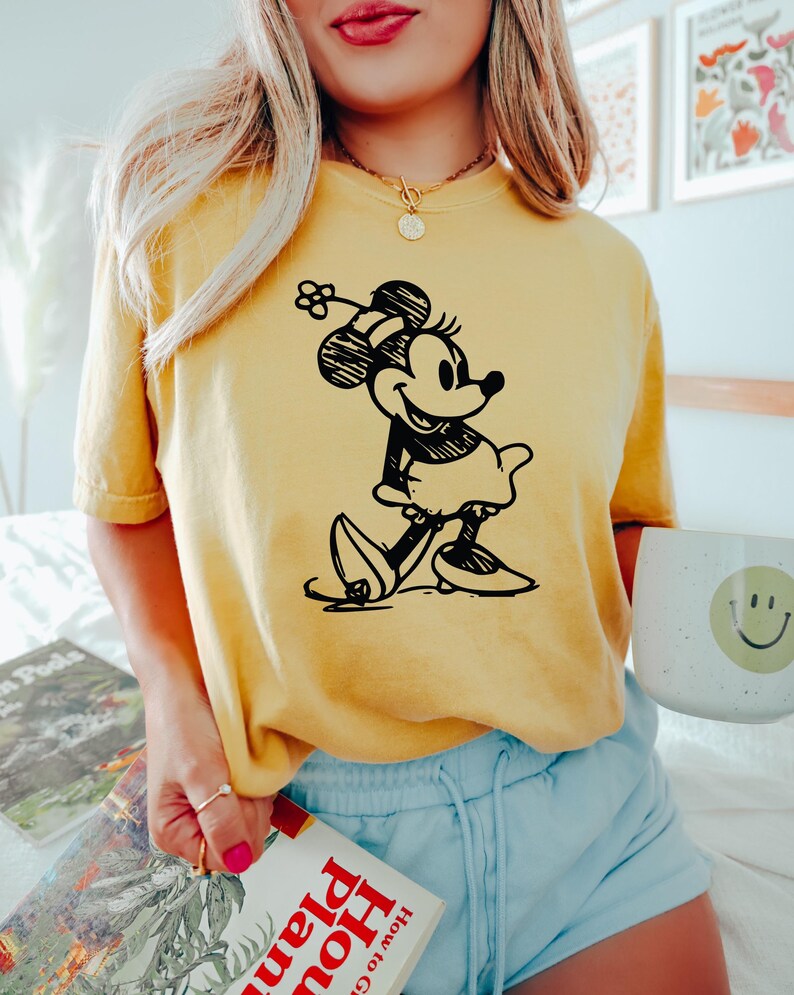 May include: Yellow t-shirt featuring a black and white illustration of Minnie Mouse. The shirt is paired with light blue shorts. A white mug with a smiley face is visible.