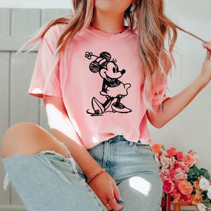 May include: A light pink t-shirt featuring a black and white illustration of Minnie Mouse. The cartoon character is depicted in a walking pose, wearing a dress and a hat with a flower. The shirt is paired with light blue jeans.