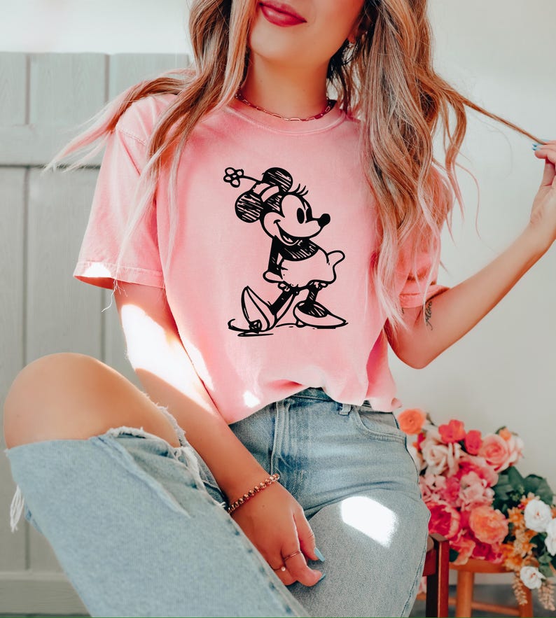 May include: A light pink t-shirt featuring a black and white illustration of Minnie Mouse. The cartoon character is depicted in a walking pose, wearing a dress and a hat with a flower. The shirt is paired with light blue jeans.