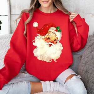 May include: Red Christmas sweatshirt with a vintage illustration of Santa Claus's face. The illustration is in a cartoon style with a white beard and a red hat with white trim.