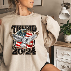 May include: A beige sweatshirt with a graphic print of an eagle flying over an American flag with the text "TRUMP 2024" in black.